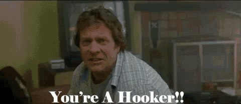 a man in a plaid shirt is standing in a living room and says `` you 're a hooker ! ''