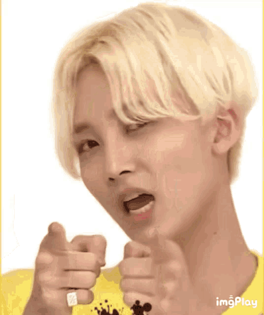 a young man with blonde hair is pointing at the camera while wearing a yellow shirt and a ring .