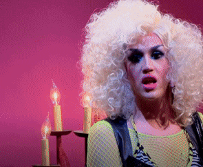 a woman in a wig stands in front of candles on a pink background