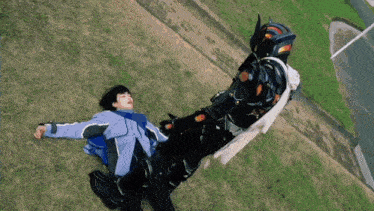 a man in a blue jacket is laying on the grass next to a man in a black costume