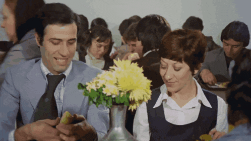 a man and a woman sit at a table with a vase of flowers in front of them