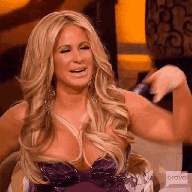 a blonde woman in a purple dress is sitting in a chair with her hand up .