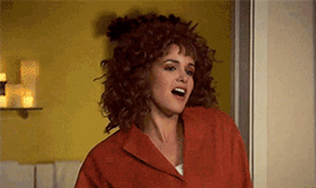 a woman with curly hair and a red jacket is standing in a room .