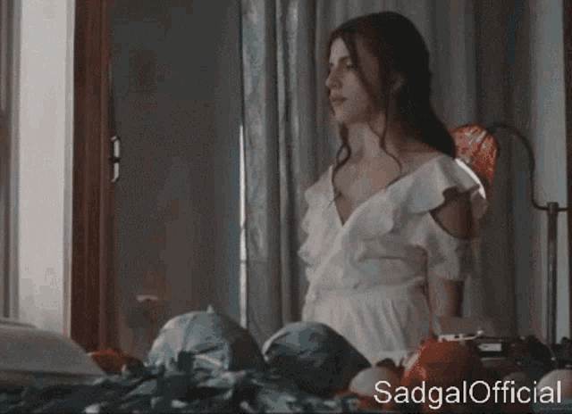 a woman in a white dress is sitting on a bed looking out the window