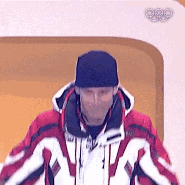 a man wearing a black hat and a red and white jacket stands in front of an olympic logo