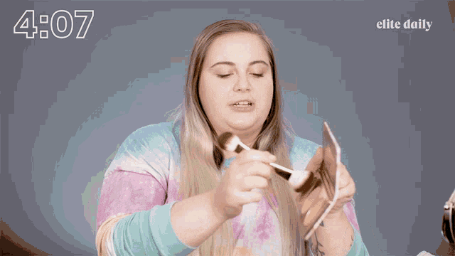 a woman in a tie dye shirt is applying makeup in front of a screen that says elite daily