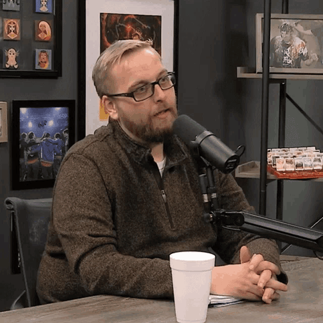 a man wearing glasses sits in front of a microphone with a cup of coffee in front of him
