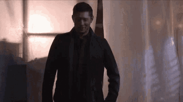 a man in a black jacket and tie is standing in a dark room with his hands in his pockets .