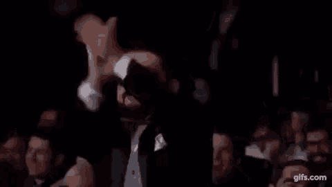 a man in a tuxedo and bow tie is raising his arms in the air in a crowd of people .