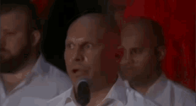 a bald man is speaking into a microphone in front of a red curtain