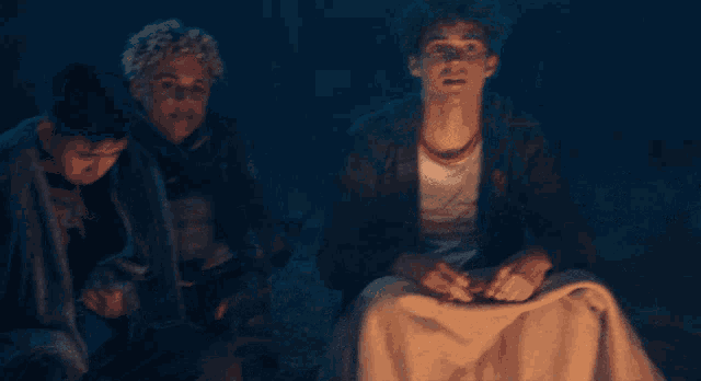three young men are sitting around a campfire with one wearing a white shirt that says california