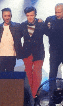 three men are standing next to each other on a stage with a fan in the foreground