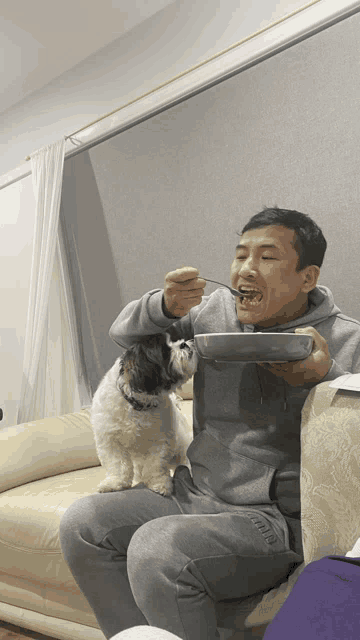 a man is sitting on a couch with a dog and eating from a bowl