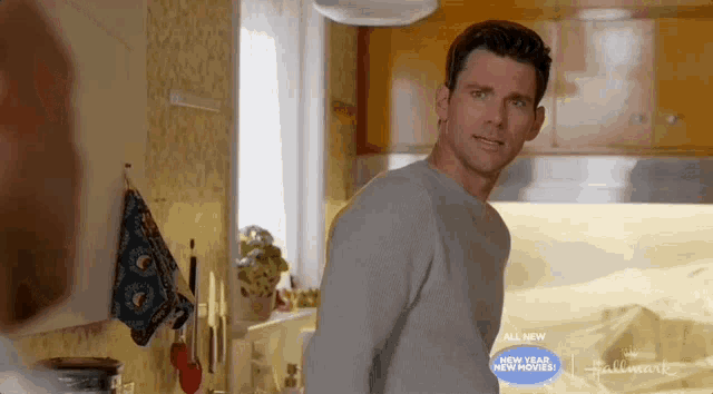 a man in a gray sweater is standing in a kitchen looking at something .