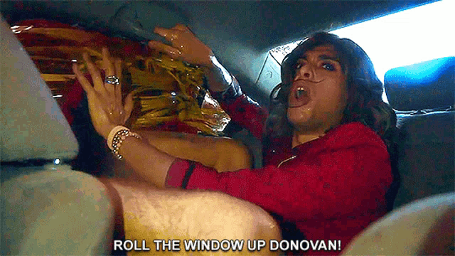 a man in a red shirt is sitting in the back seat of a car and says roll the window up donovan