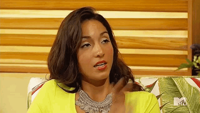 a woman wearing a yellow top and a silver necklace is sitting on a couch with a mtv logo in the background .