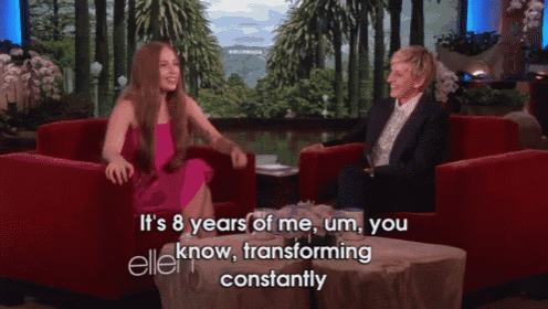 a woman in a pink dress sits next to an ellen degeneres on a red couch