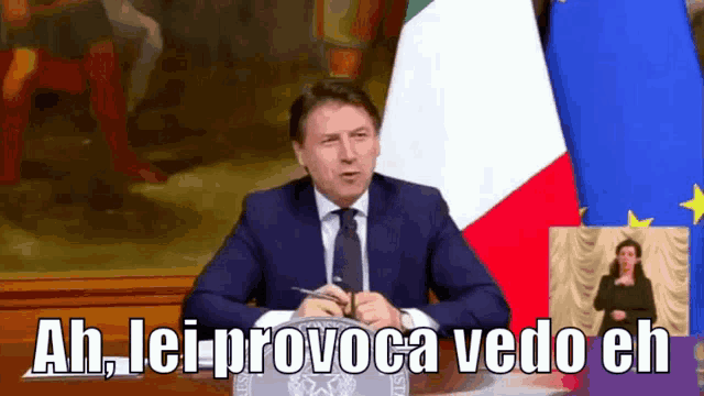 a man in a suit and tie is giving a speech while sitting at a podium in front of an italian flag .