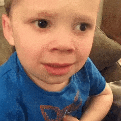a little boy in a blue shirt is sitting on a couch making a funny face .