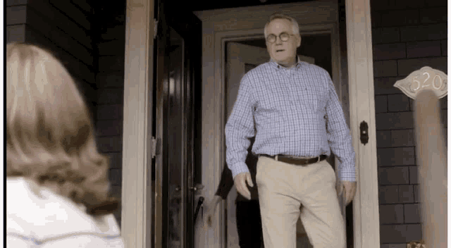 a man in a plaid shirt and khaki pants is standing in front of a door with the number 520 written on it .