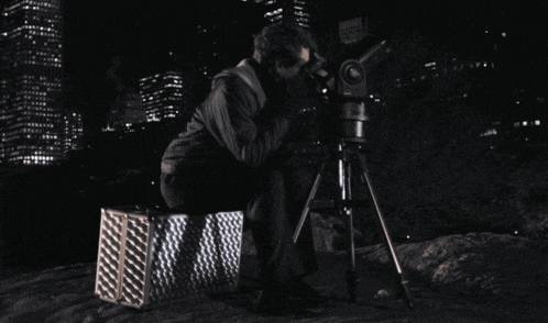 a man is looking through a telescope while sitting on a box