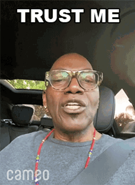 a man wearing glasses and a necklace is sitting in a car with the words trust me above him