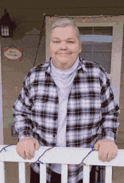 a man in a plaid shirt stands on a porch with a merry christmas sign above him
