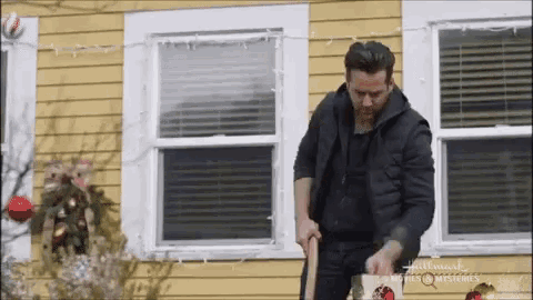 a man in a black jacket is standing in front of a yellow house holding a shovel .