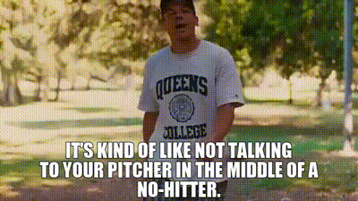 a man wearing a queens college shirt is standing in a park with trees in the background .