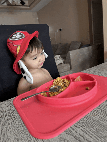 a baby wearing a fireman hat is eating from a red tray