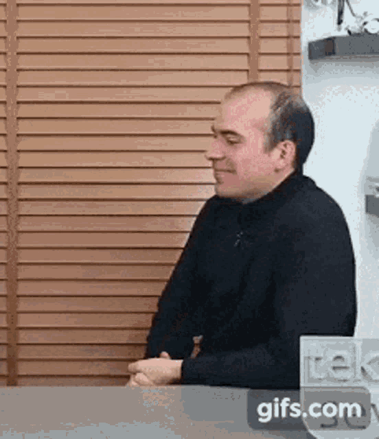 a man is sitting at a desk in front of a window with blinds and a sign that says trek
