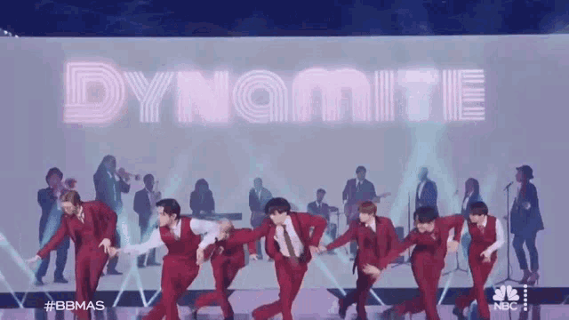 a group of men in red suits are dancing in front of a large screen that says dynamite