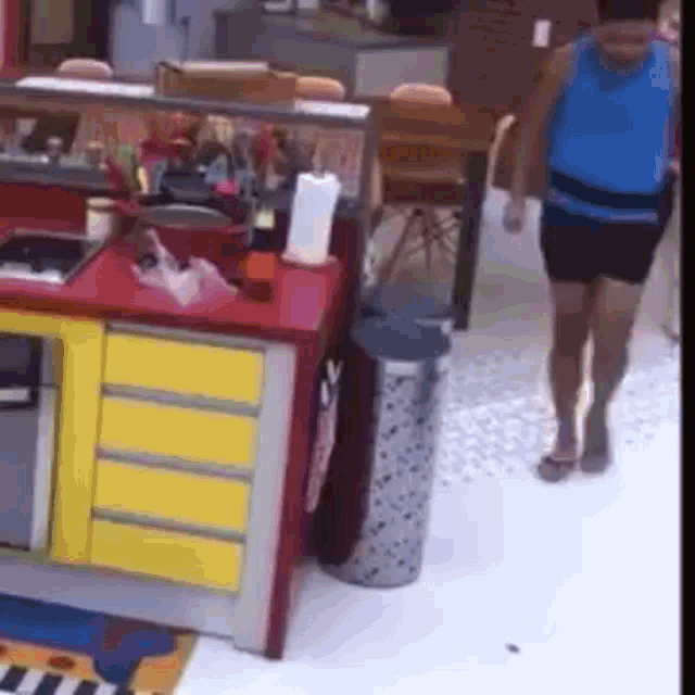a woman in a blue tank top is walking in a kitchen with a trash can .