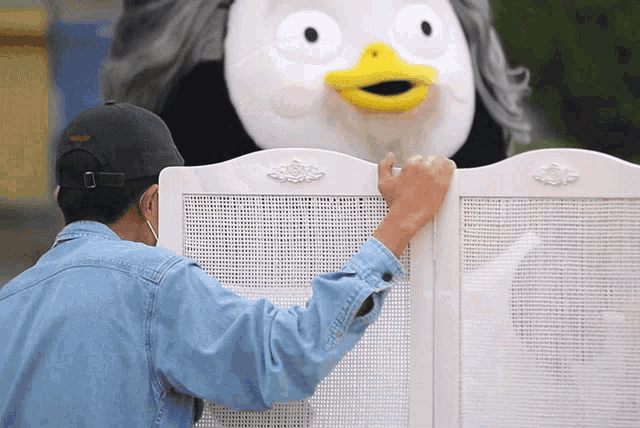 a man in a denim shirt is touching a white fence with a stuffed animal behind it