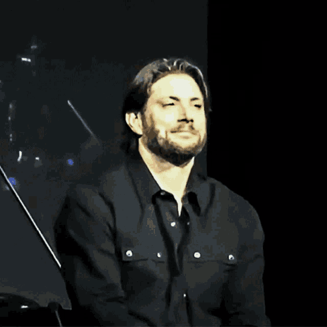 a man with a beard wearing a black shirt is sitting on a stage