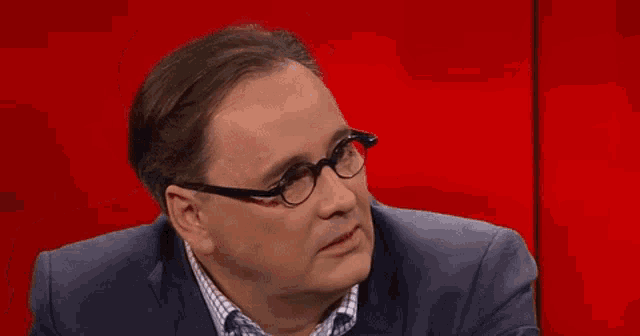 a man wearing glasses and a suit is sitting on a red background