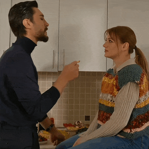 a man and a woman are sitting in a kitchen and the man is pointing at the woman 's face