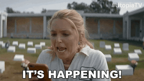 a woman is holding a cell phone in front of a cemetery and saying `` it 's happening '' .