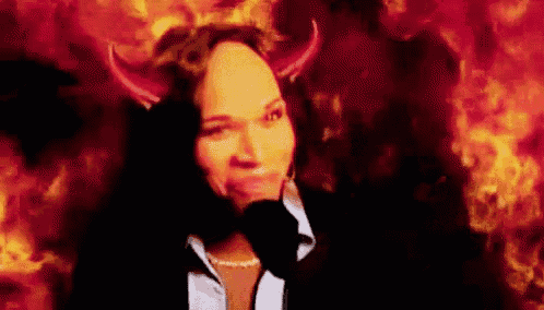 a woman in a suit and tie with devil horns on her head is standing in front of a fire .