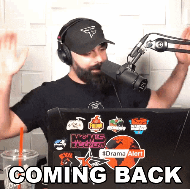 a man wearing headphones and a hat stands in front of a microphone and a laptop that says coming back on it