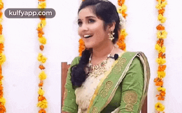 a woman in a green and white saree is sitting in a chair with flowers behind her .