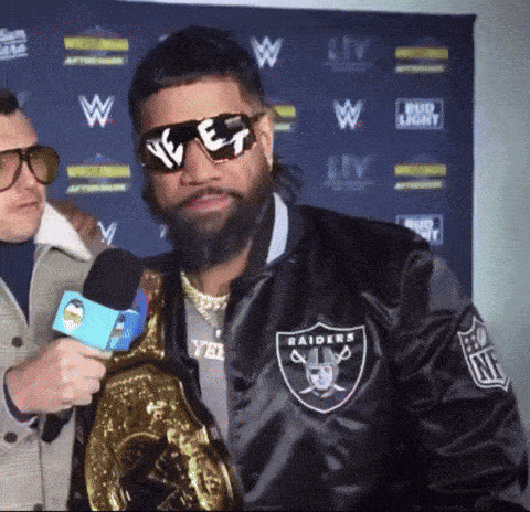 a man wearing sunglasses and a raiders jacket is holding a microphone