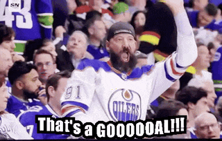 a man with a beard is sitting in the stands watching a hockey game and raising his fist in the air .