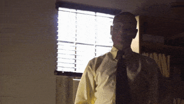a man in a yellow shirt and black tie stands in front of a window with blinds