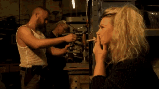 a woman is smoking a cigarette while a man works on a machine in the background