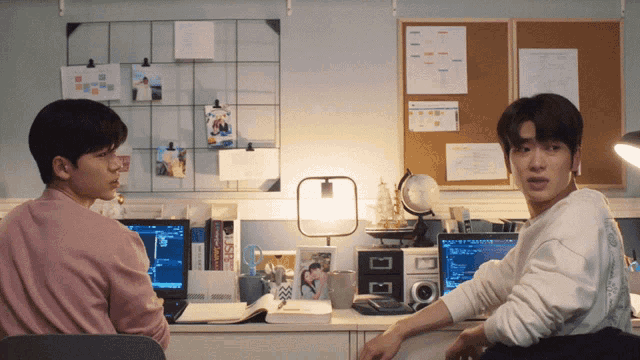 two men are sitting at a desk with computers and a bulletin board behind them