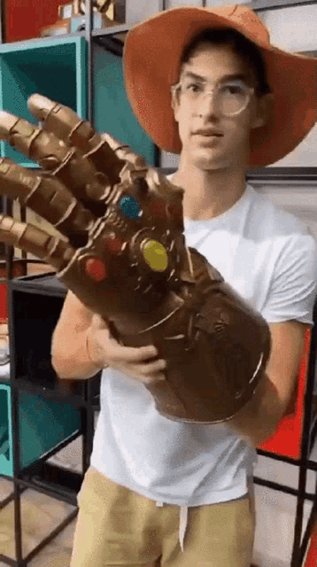a young man wearing a hat and glasses is holding a giant infinity gauntlet .