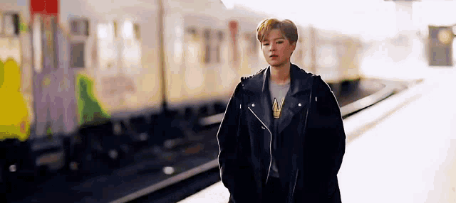a woman in a black leather jacket is standing on a train platform next to a train .