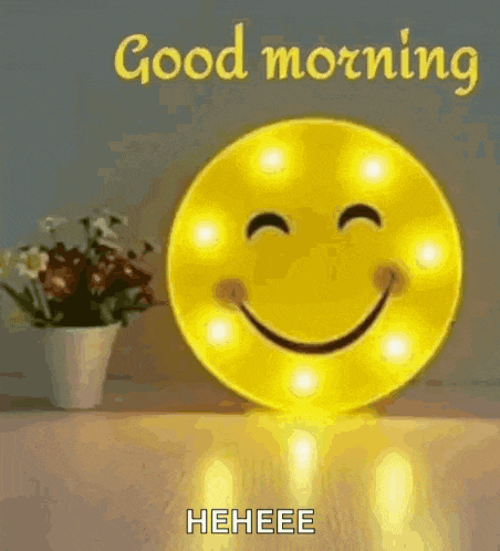 a smiley face is sitting on a table next to a vase of flowers and a good morning message .