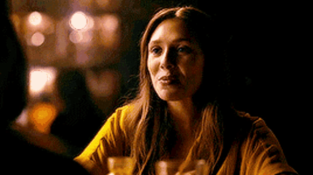a woman in a yellow shirt is sitting at a table in a dark room with a drink in her hand .